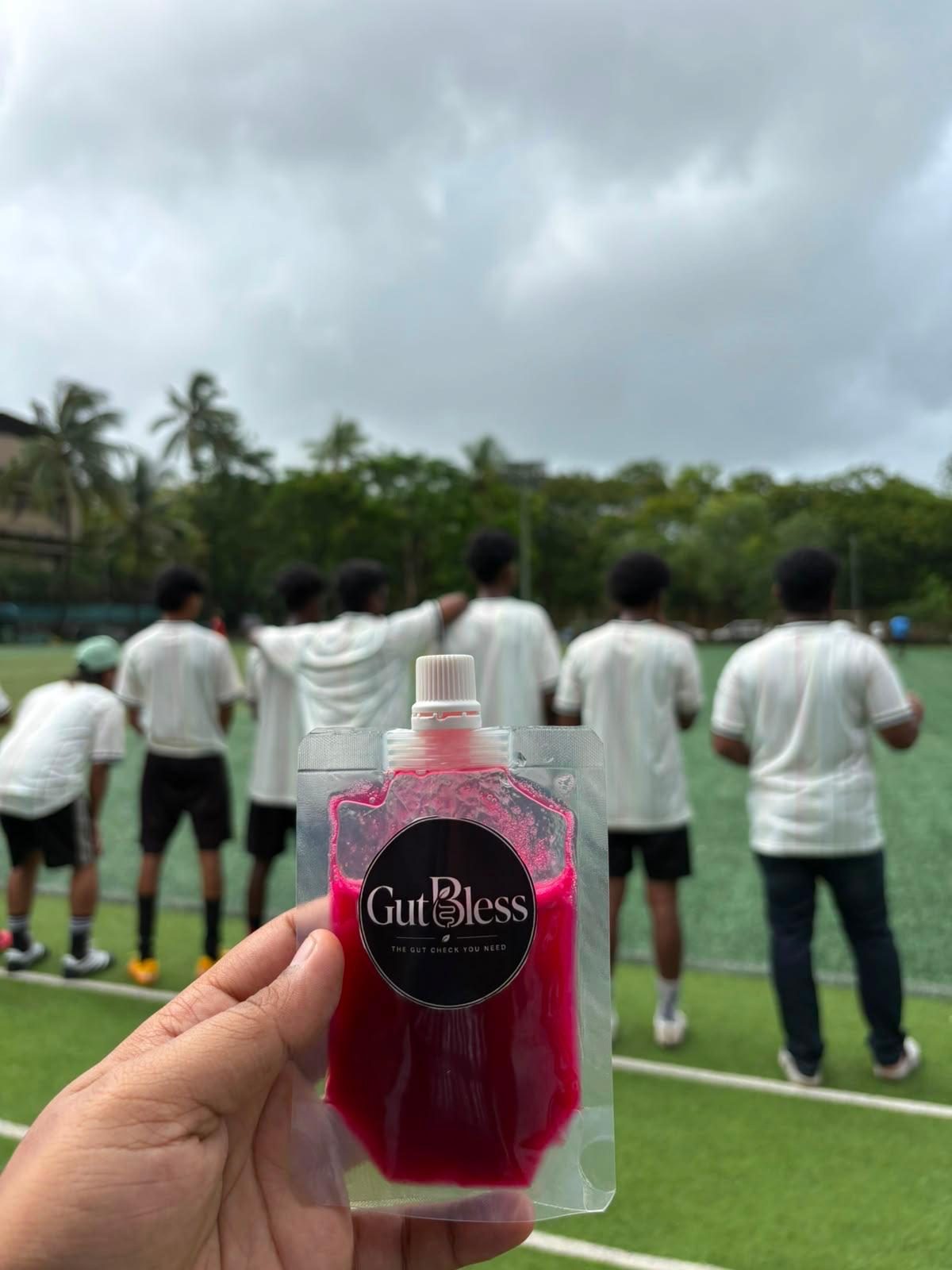 A GutBless cold-pressed juice pouch held up in front of footballers lined up on a turf ground